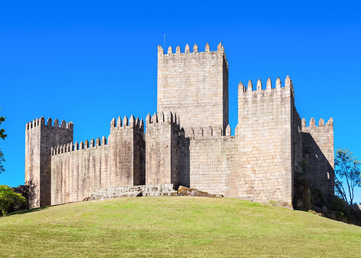 A stone medieval castle with crenellated walls and towers sits on a grassy hill under a clear blue sky.