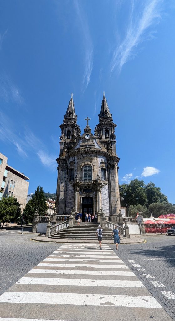 A historical church with twin spires under a blue sky, viewed from across a crosswalk. Several people are near the entrance, and trees and outdoor seating are visible on the right.