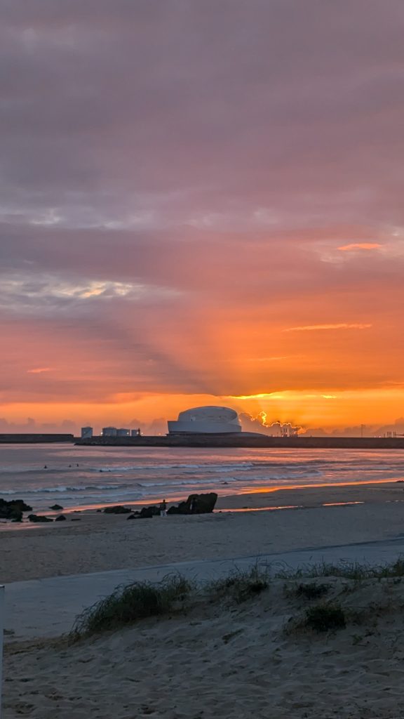 A sunset over an industrial facility with smokestacks emitting smoke, silhouetted against the horizon. A beach with waves is in the foreground.