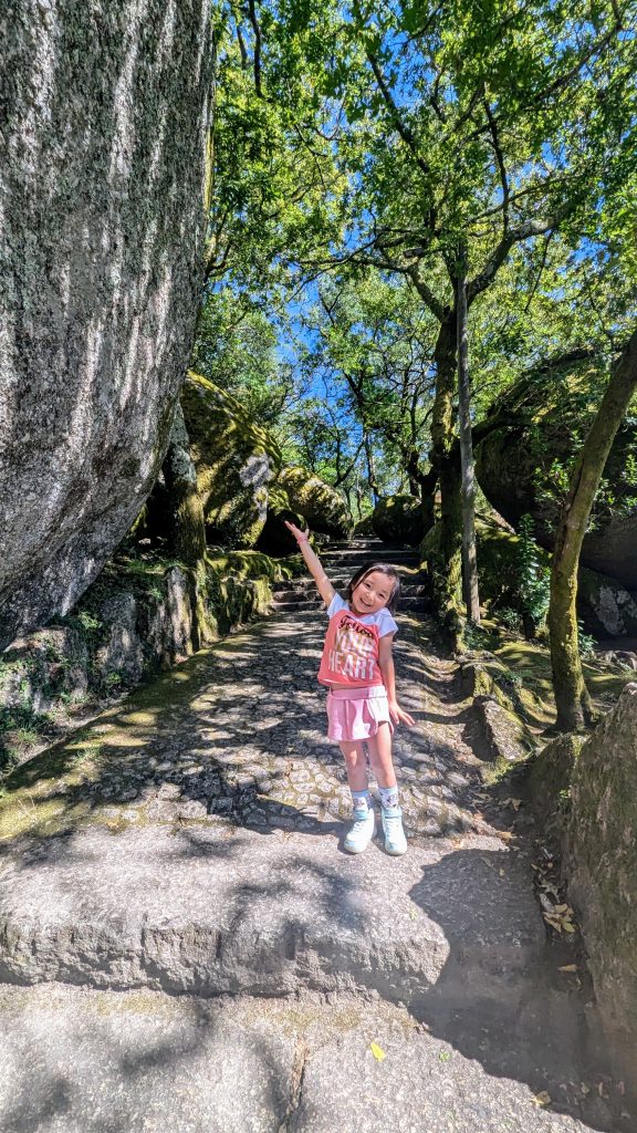 A child stands on a stone pathway surrounded by large rocks and trees, smiling and pointing upwards with one arm.