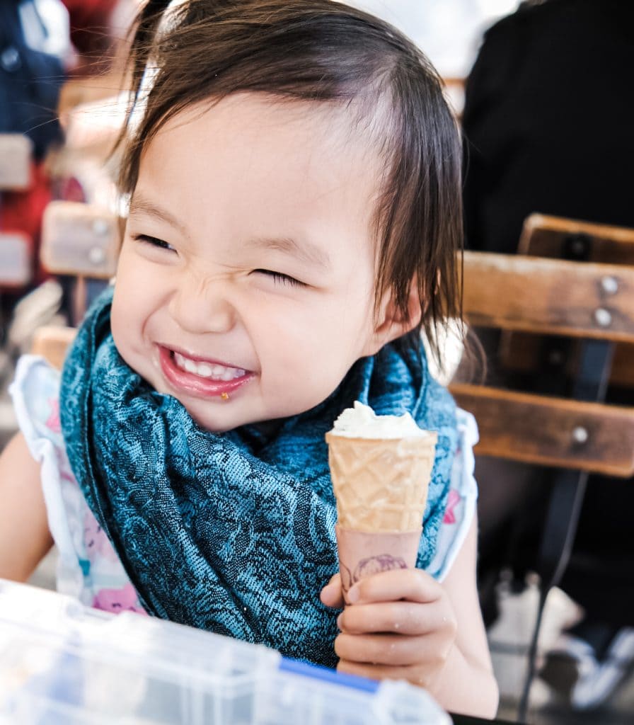 A young child with a blue scarf smiles broadly while holding an ice cream cone in an outdoor setting.