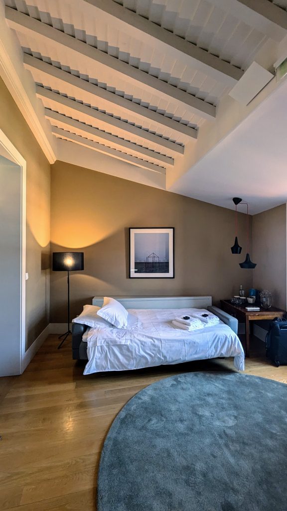 A hotel room with wooden flooring, a bed with white sheets, a floor lamp, a desk with two pendant lights, and an angled ceiling with exposed beams. A framed picture and various items are on the desk.