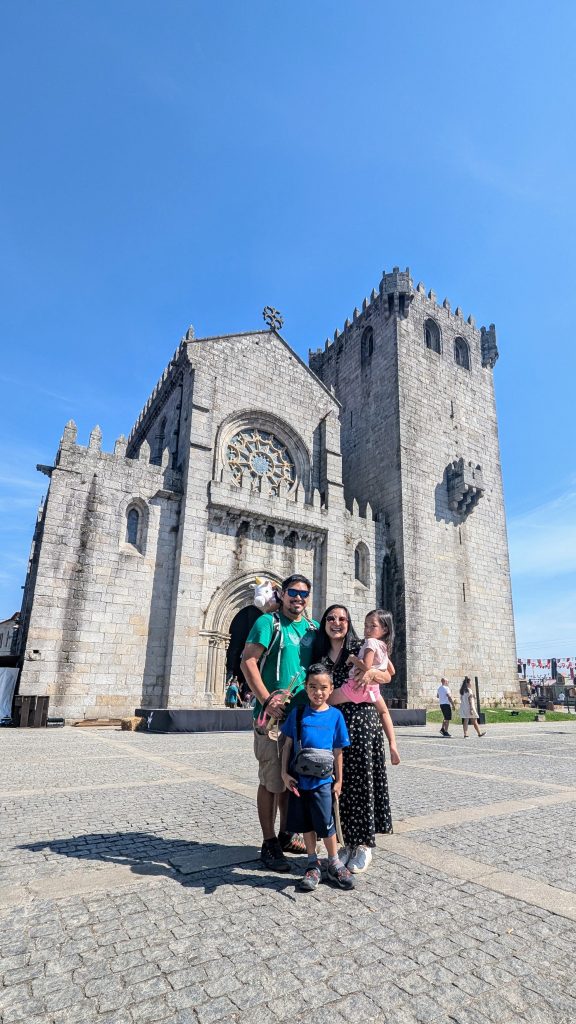 A family of four stands in front of a historic stone cathedral on a sunny day. The parents are holding hands with their young children.