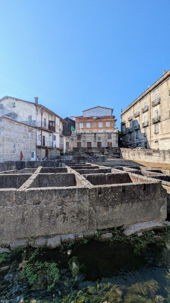 Wide-angle view of an old stone structure with rectangular pits in front of historic buildings under a clear blue sky.