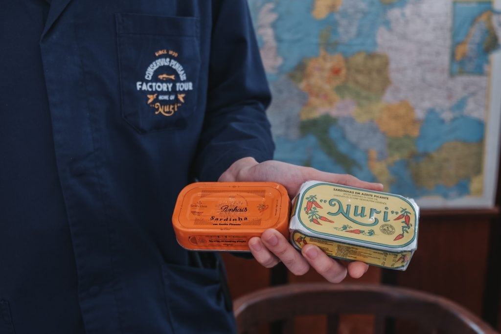 Person holding two tins of sardines, one orange and one white, with "Conservas Pinhais" on their blue shirt, and a map in the background.