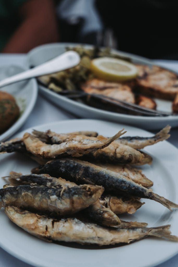A plate of fried sardines with a side dish in the background containing grilled vegetables and lemon slices.