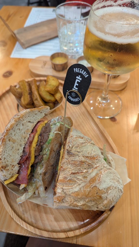 A sandwich with meat, cheese, and vegetables on a wooden tray, accompanied by potato wedges and a glass of beer. A toothpick with a tag is inserted in the sandwich.