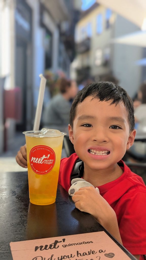 A young boy in a red shirt sits at a table, holding a drink with a straw in front of him. The drink has a label that says "nut' Guimarães." The background shows a blurred, busy cafe or restaurant setting.