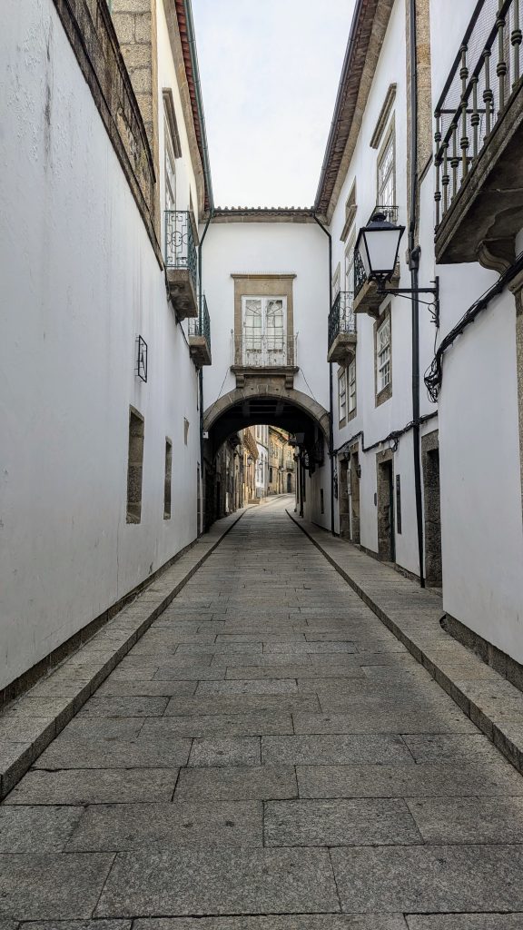 A narrow cobblestone street flanked by white buildings with balconies. The buildings form an archway overhead, leading to a distant point. It appears to be a quiet, historic European street.