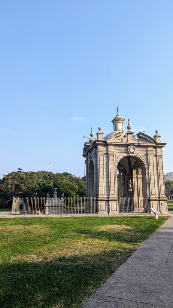 A stone pavilion with classical architectural elements stands on a grassy area under a clear blue sky, surrounded by a metal fence. Trees and pathways are visible in the background.