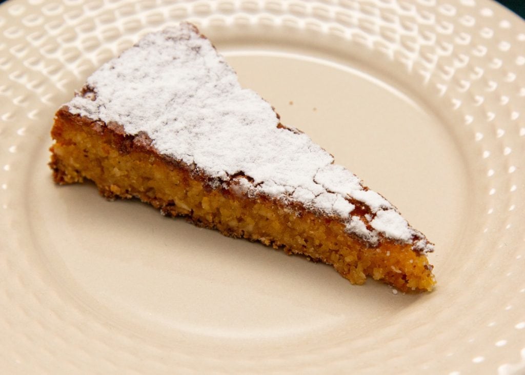 A slice of almond cake dusted with powdered sugar sits on a beige plate with a textured rim.