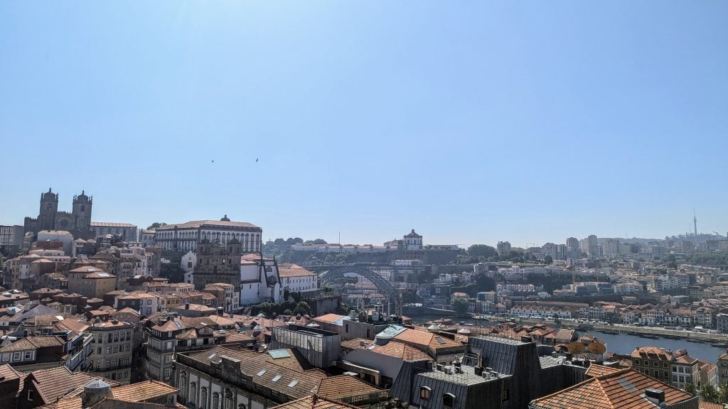 Panoramic view of Porto, Portugal, featuring the Douro River and the Dom Luís I Bridge, with historic buildings and red-tiled roofs under a clear blue sky.