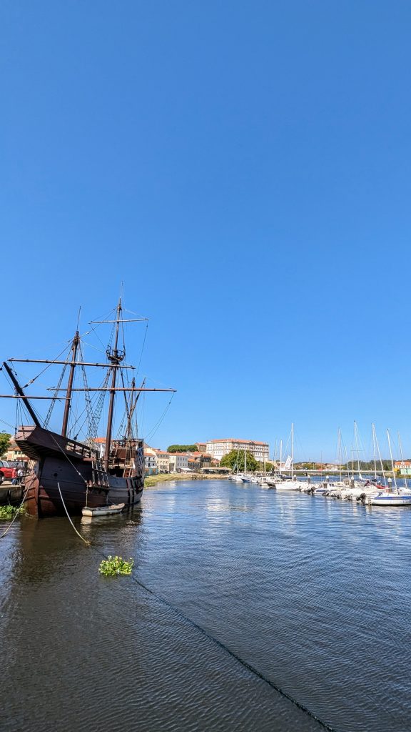 A historic wooden ship docked next to modern sailboats on a calm river under a clear blue sky, with buildings in the background.