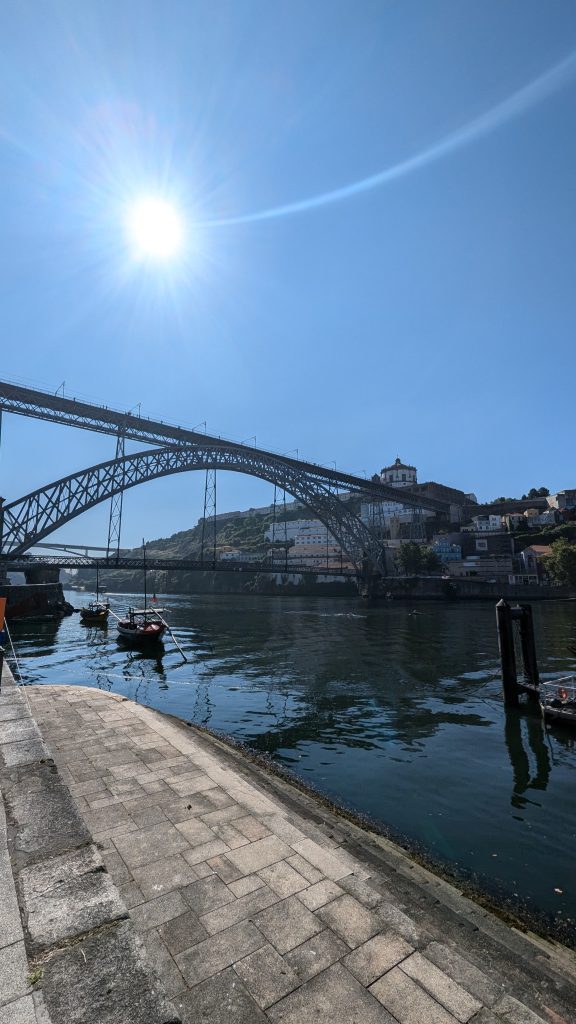 Sun shining over a large bridge spanning a river, with small boats below. A paved path runs alongside the water, leading toward distant hills and buildings.