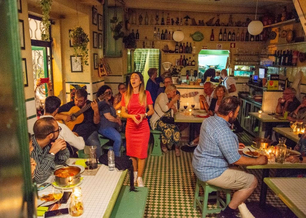 People enjoy a lively evening in a cozy restaurant. A woman in a red dress sings fado while a guitarist plays. Diners sit at tables with food and drinks, surrounded by warm lighting and decorations.