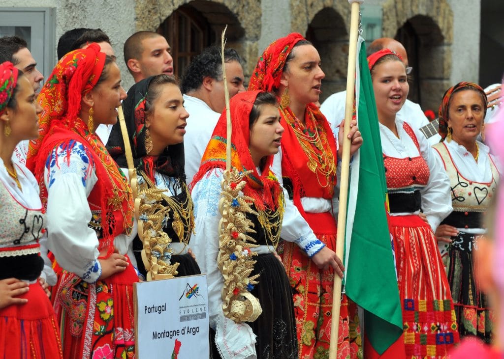 A group of people in traditional Portuguese attire, with colorful red and white costumes, stand together. One person holds a Portuguese flag.