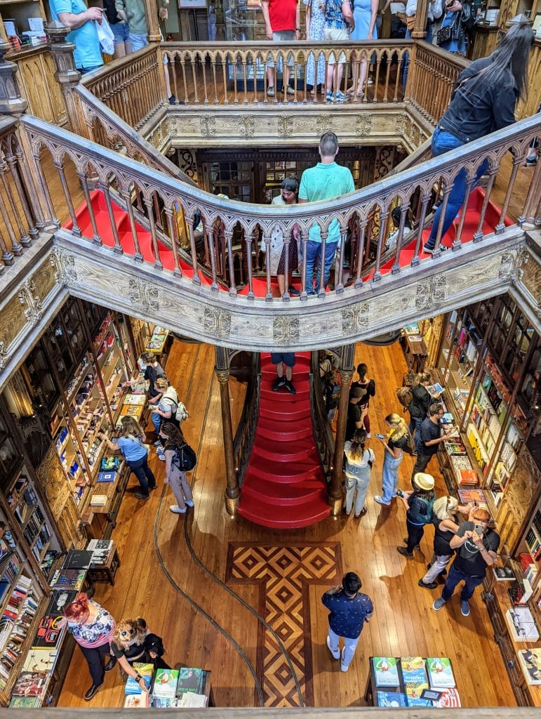 Interior of a grand multi-level bookstore with ornate wooden railings, red carpeted stairs, and people browsing books on both floors.