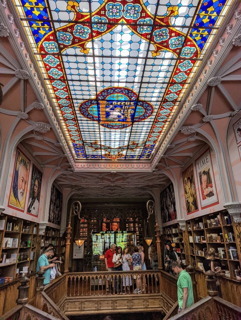 A colorful, ornate stained glass ceiling above a wooden staircase in a bookstore, with people browsing and framed magazines on the walls.