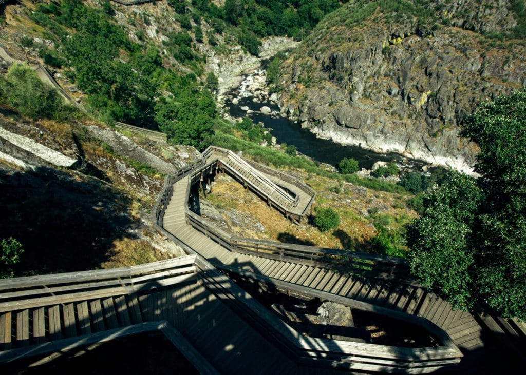 A wooden staircase zigzags down a hillside with rocky terrain and greenery, leading to a river below.