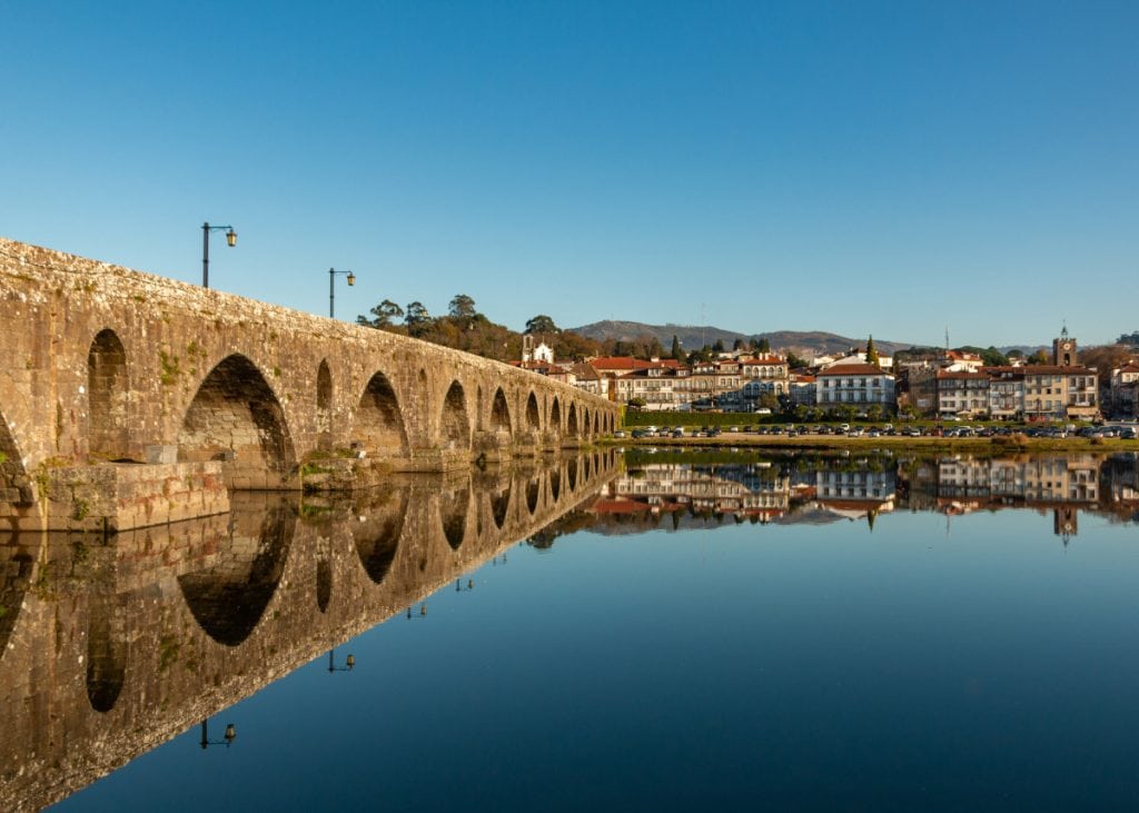 A historic stone bridge with arches is reflected in a calm river. The background features a town with white buildings under a clear blue sky.