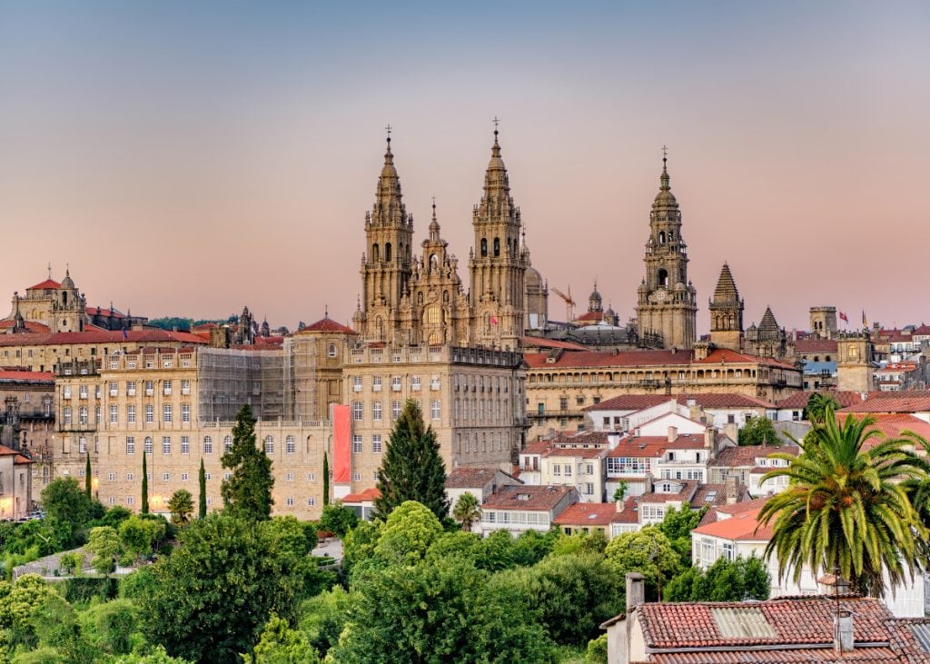 Cityscape of Santiago de Compostela at sunset with its iconic cathedral spires, surrounded by historic buildings and lush greenery.