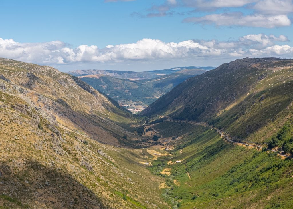 View of a wide valley between two green and brown mountains under a partly cloudy sky, with a distant town visible at the bottom.