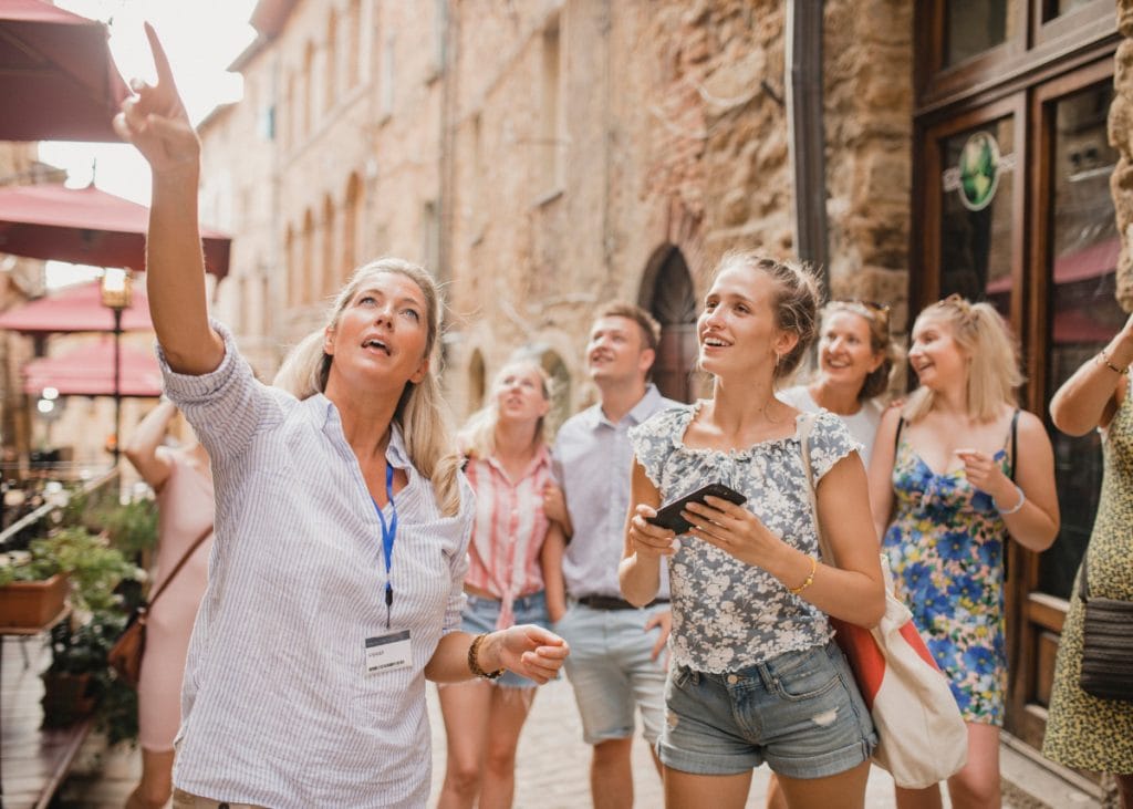 A group of people on a guided tour in a historic area with stone buildings. A woman gestures upward while others look in the same direction. Several are smiling, and one holds a phone