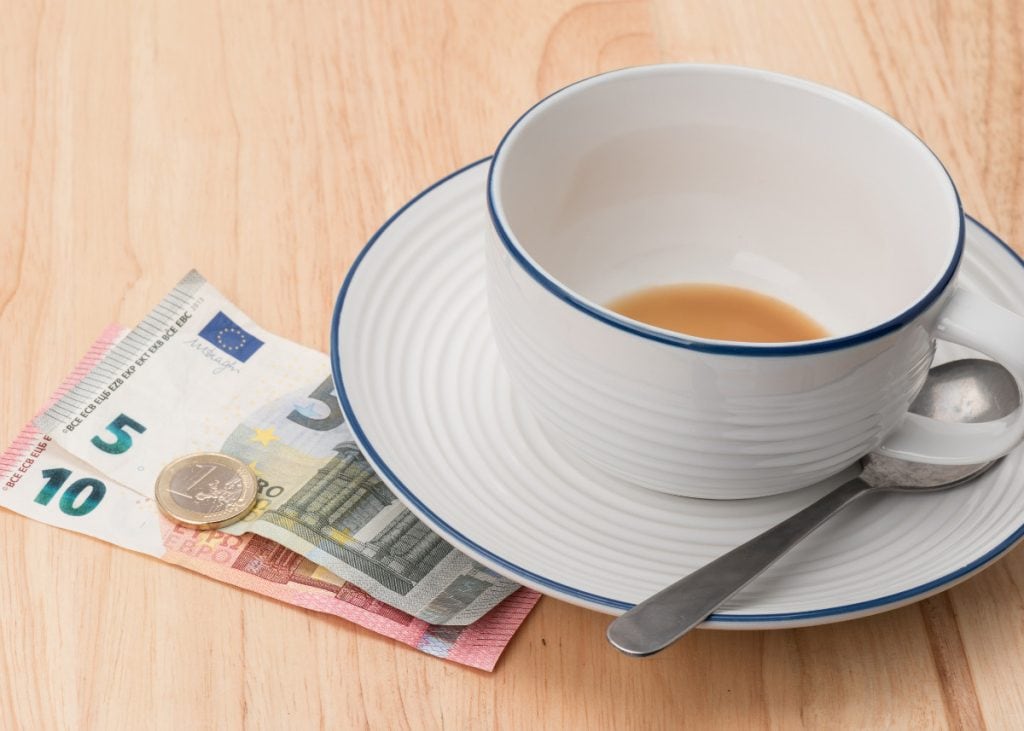 A white cup with a saucer holding a small amount of coffee, a metal spoon, and Euro banknotes and a coin on a wooden surface.