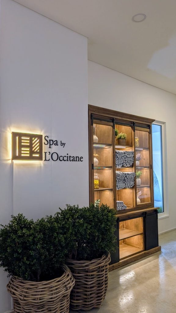 Lobby of a spa with a wooden display shelf featuring neatly arranged towels and plants, next to a sign that reads "Spa by L'Occitane".