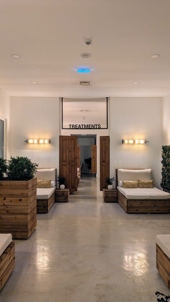 Spa waiting area with wooden seating, white cushions, potted plants, and ambient lighting. A sign above the doors reads "TREATMENTS.