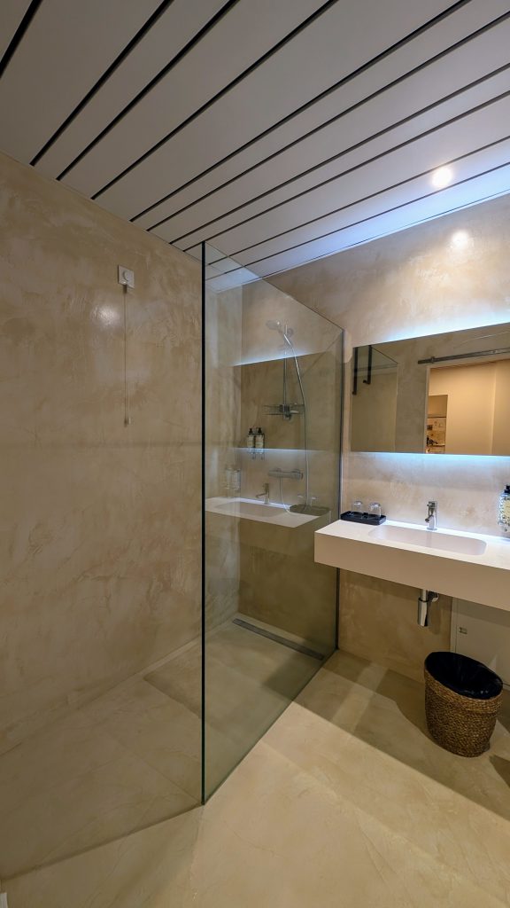 Modern bathroom with beige walls, a walk-in glass shower, a rectangular white sink with silver faucet, large mirror, and a brown wicker trash bin on the floor.