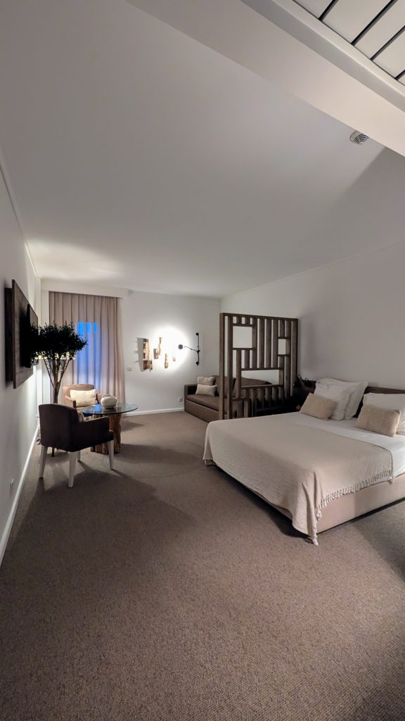 Spacious hotel room with a double bed, sofa, bunk bed, small table, and wall-mounted TV. Neutral colors and modern decor create a cozy atmosphere.