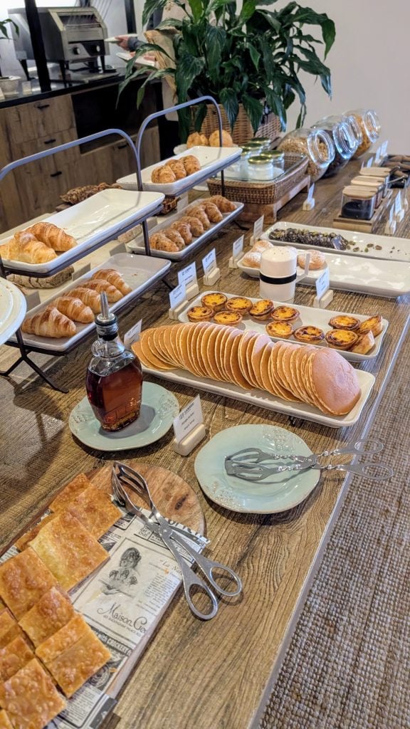 A display of breakfast items on a wooden table, including pancakes, pastries, croissants, and a jar of syrup, with plants in the background.
