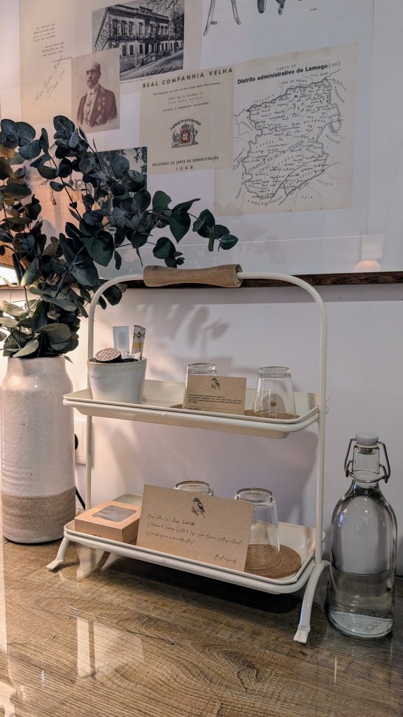 A two-tiered metal stand holds glasses and coasters, flanked by a large vase with eucalyptus. Wall art includes old documents and a map, with a bottled drink on a wooden surface.