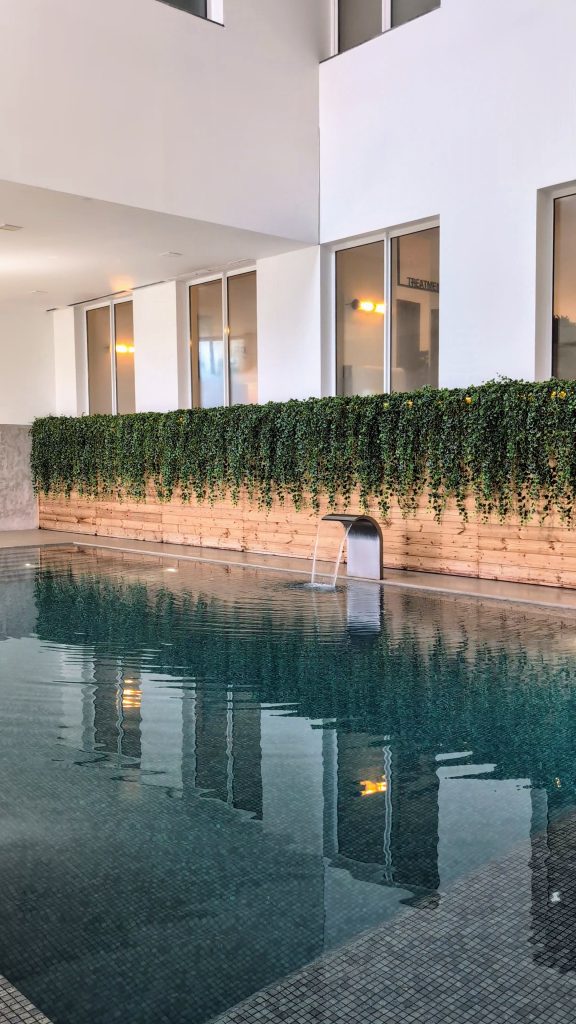 Indoor swimming pool with clear water, surrounded by white walls and windows. A wooden planter with greenery hangs above the pool ladder.