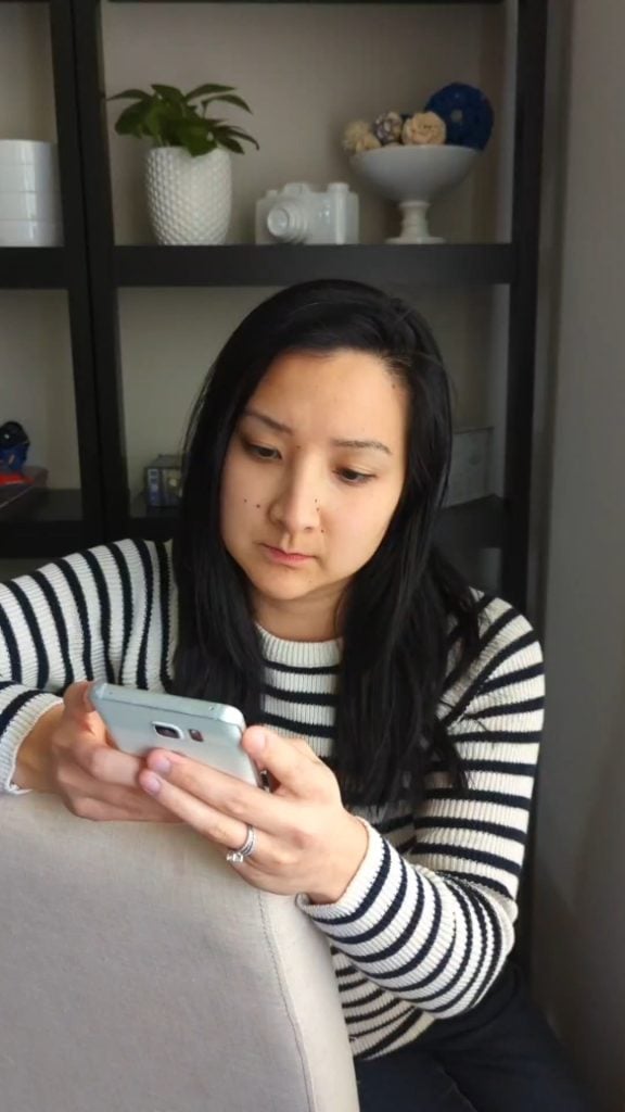 A woman with long dark hair wearing a black and white striped sweater sits indoors, looking at her smartphone. Shelves with decorative items are in the background.