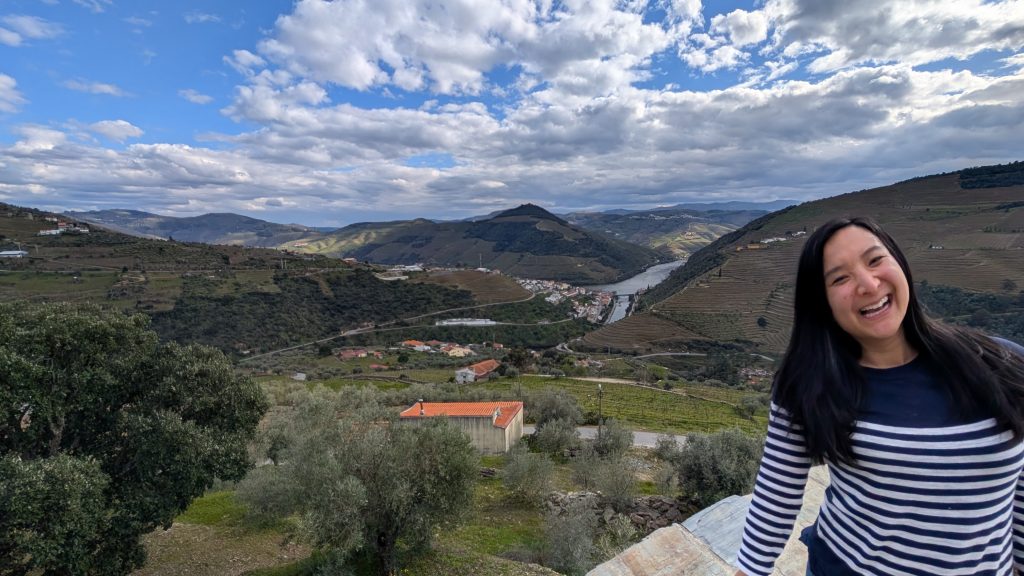 A woman in a striped shirt stands smiling in the foreground with the Douro Valley in the background.