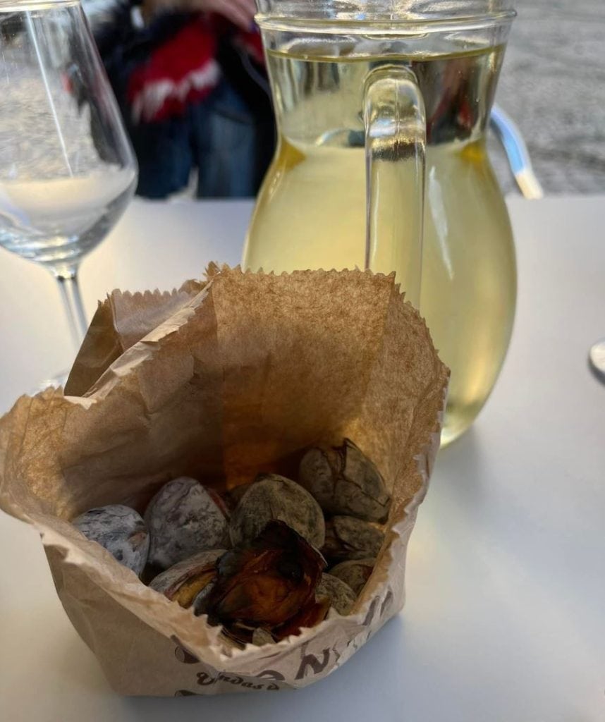 A brown paper bag of roasted chestnuts sits on a table next to a glass pitcher of white wine and an empty wine glass.