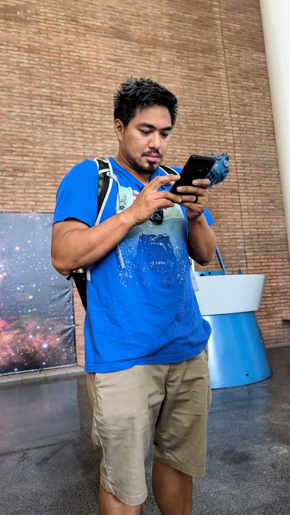 A man in a blue t-shirt and khaki shorts stands indoors, looking at his phone. He has a backpack and there is a brick wall and a space-themed display in the background.