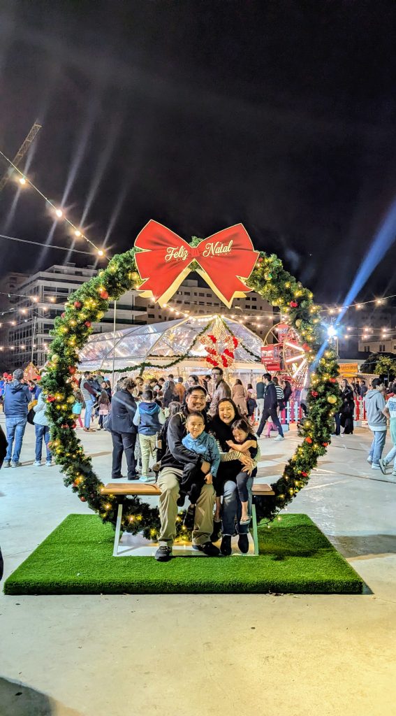 A family of four sits on a bench inside a large Christmas wreath with a “Feliz Natal” bow at a festive outdoor event, with lights and people in the background at night.