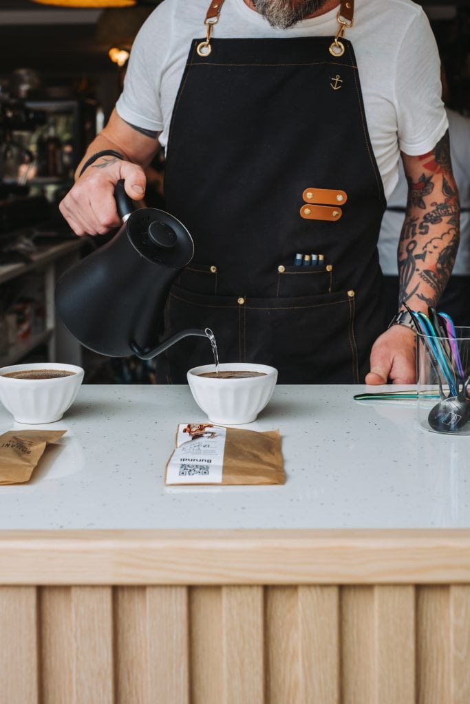 A person wearing an apron pours hot water from a kettle into a coffee dripper over a cup on a counter, with coffee equipment and a bag of beans nearby.