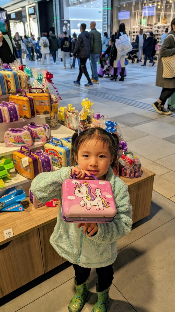 A young child stands in a shopping mall holding a pink lunchbox with a unicorn design, with a display of similar lunchboxes and shoppers in the background.