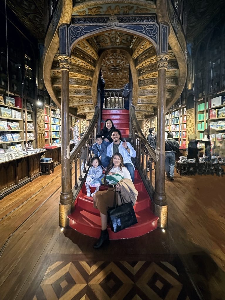 A family of five poses on red steps beneath an ornate wooden spiral staircase inside a library or bookstore with shelves of books on either side.
