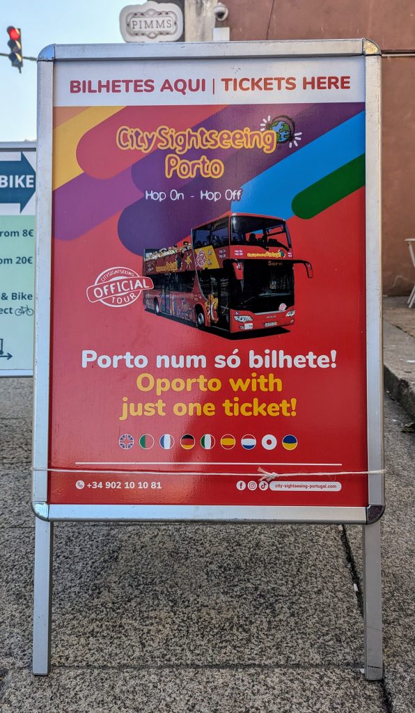 A sidewalk sign advertises City Sightseeing Porto tours with an image of a red double-decker bus and mentions tickets available in multiple languages.
