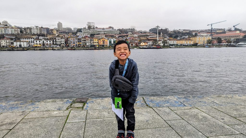 A young boy stands on a riverside promenade, smiling widely at the camera. A cityscape with colorful buildings and cranes is visible across the water under a gray sky.