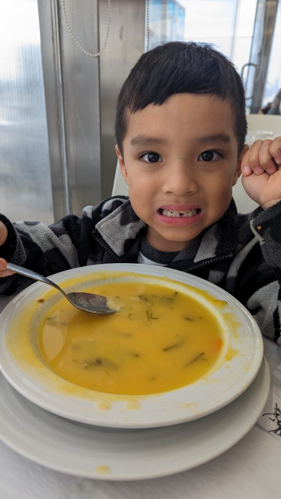 A young boy sits at a table holding a spoon while looking at a bowl of caldo verde, bright yellow in color, in front of him.