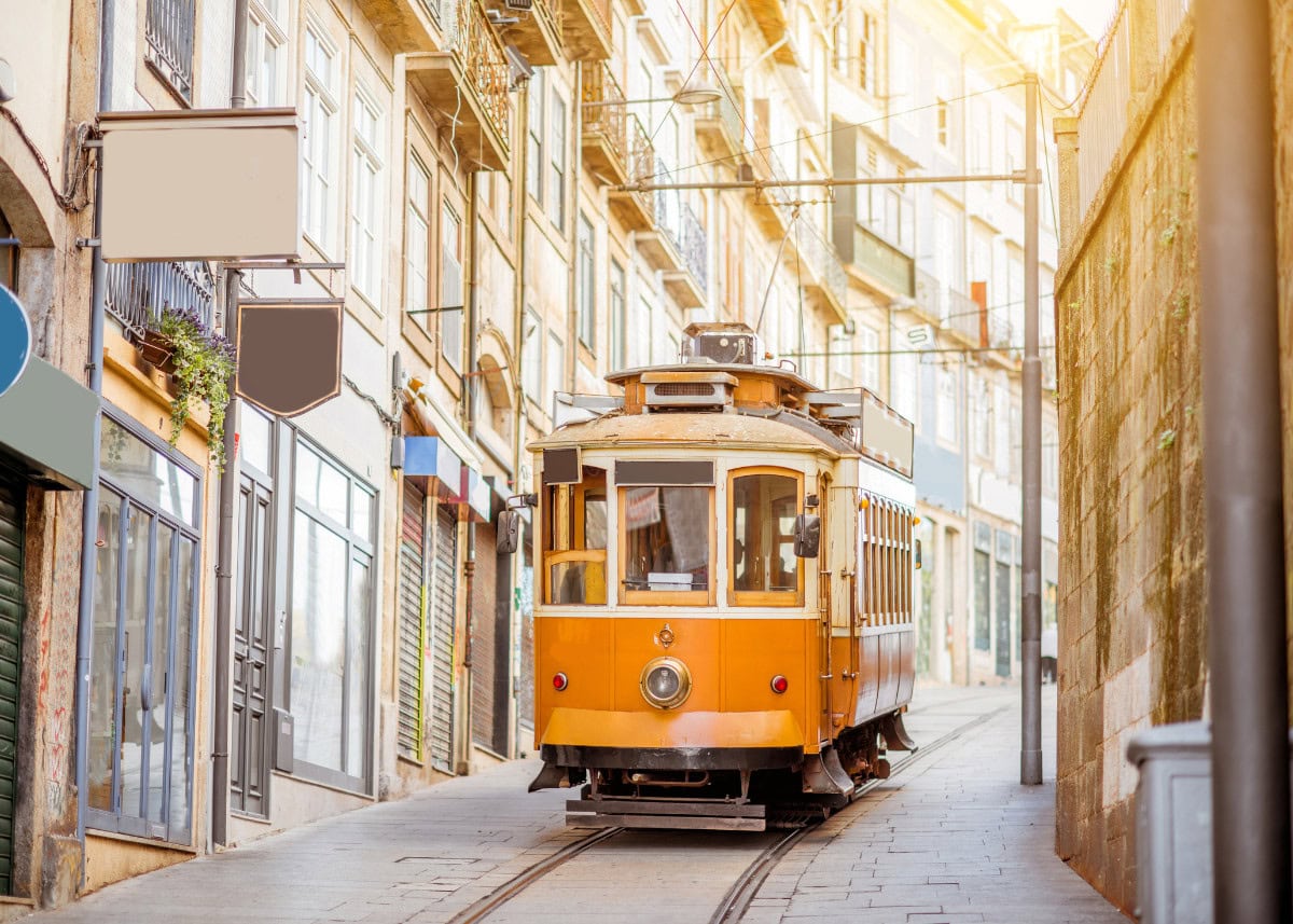Yellow tram travels along narrow, cobblestone street lined with old buildings under bright sunlight.