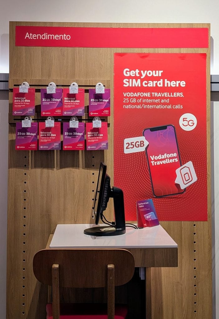 Display stand with Vodafone Travellers SIM cards and a sign promoting 25GB of internet and national/international calls, next to a computer and a small wooden table.