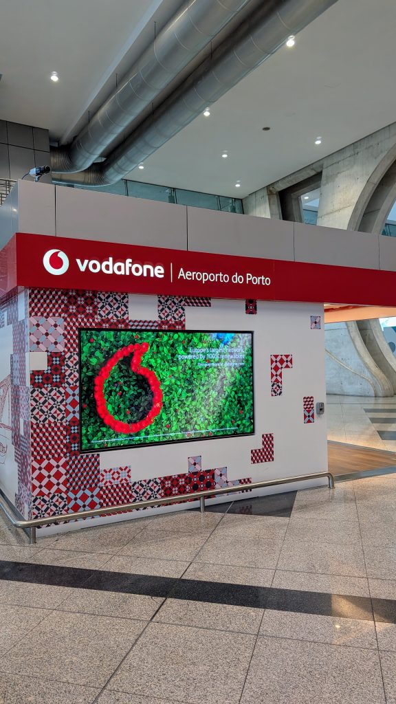 Vodafone kiosk at Porto Airport with branded signage, a digital screen displaying the Vodafone logo, and a patterned design on the booth exterior.