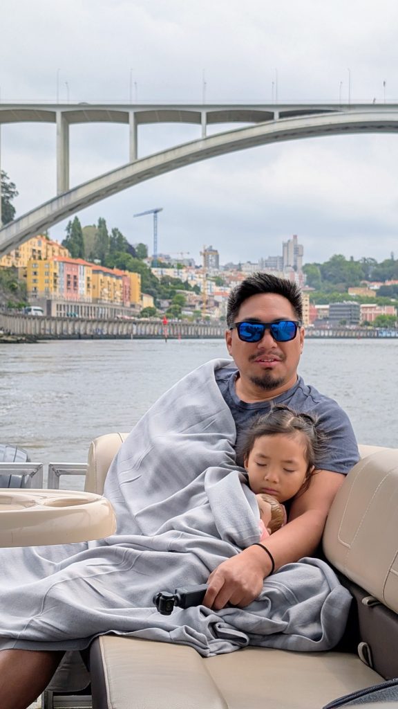 A man wearing sunglasses sits on a boat, holding a sleeping child wrapped in a blanket, with a cityscape and large bridge in the background.